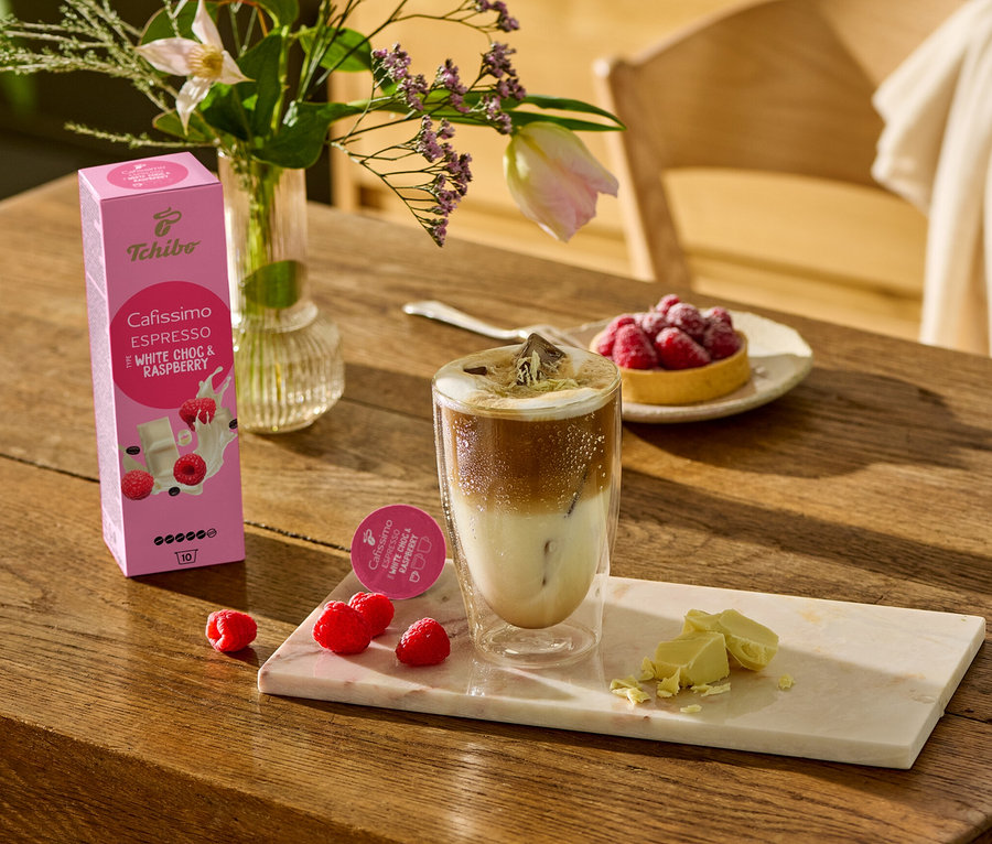 Sur une table en bois, un verre de café, des morceaux de chocolat blanc et des framboises sont posés sur une planche de marbre. À l'arrière-plan, on peut voir un paquet de Cafissimo Flavoured Espresso – White Choc & Raspberry – 10 capsules.