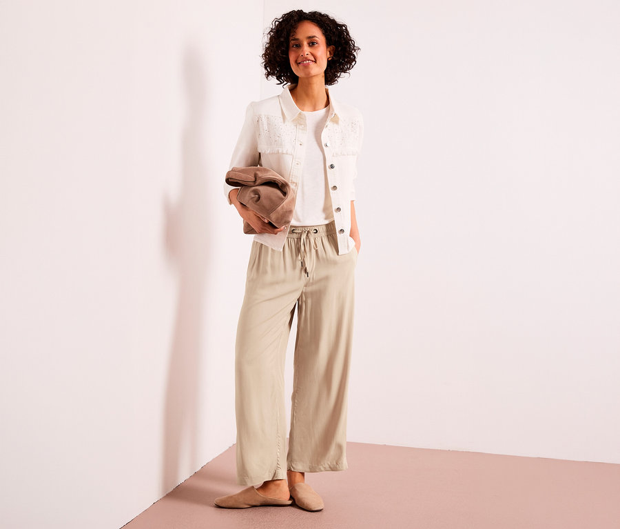 Femme souriante debout, portant un t-shirt blanc, une veste blanche et un culotte 7/8 Street One beige, tenant une pochette marron.