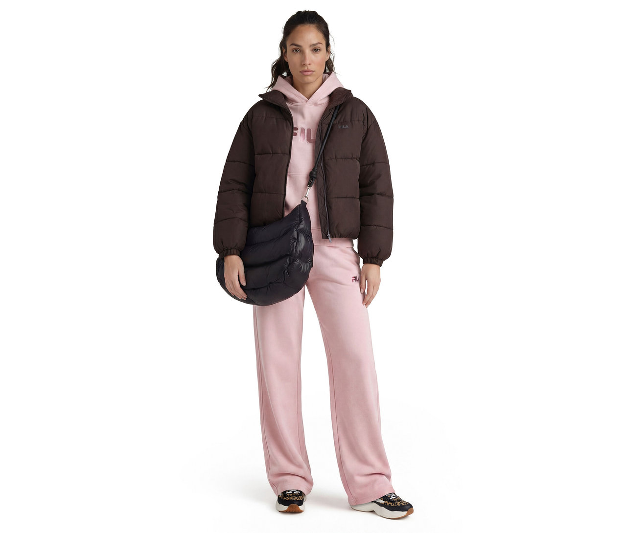 Une femme debout de face, porte une doudoune marron foncé, un sweat à capuche rose clair, un pantalon de survêtement rose clair à jambes larges et des baskets noires et imprimé léopard. Elle porte un sac bandoulière matelassé noir.