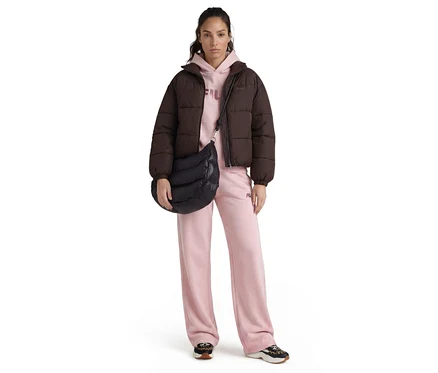Une femme debout de face, porte une doudoune marron foncé, un sweat à capuche rose clair, un pantalon de survêtement rose clair à jambes larges et des baskets noires et imprimé léopard. Elle porte un sac bandoulière matelassé noir.