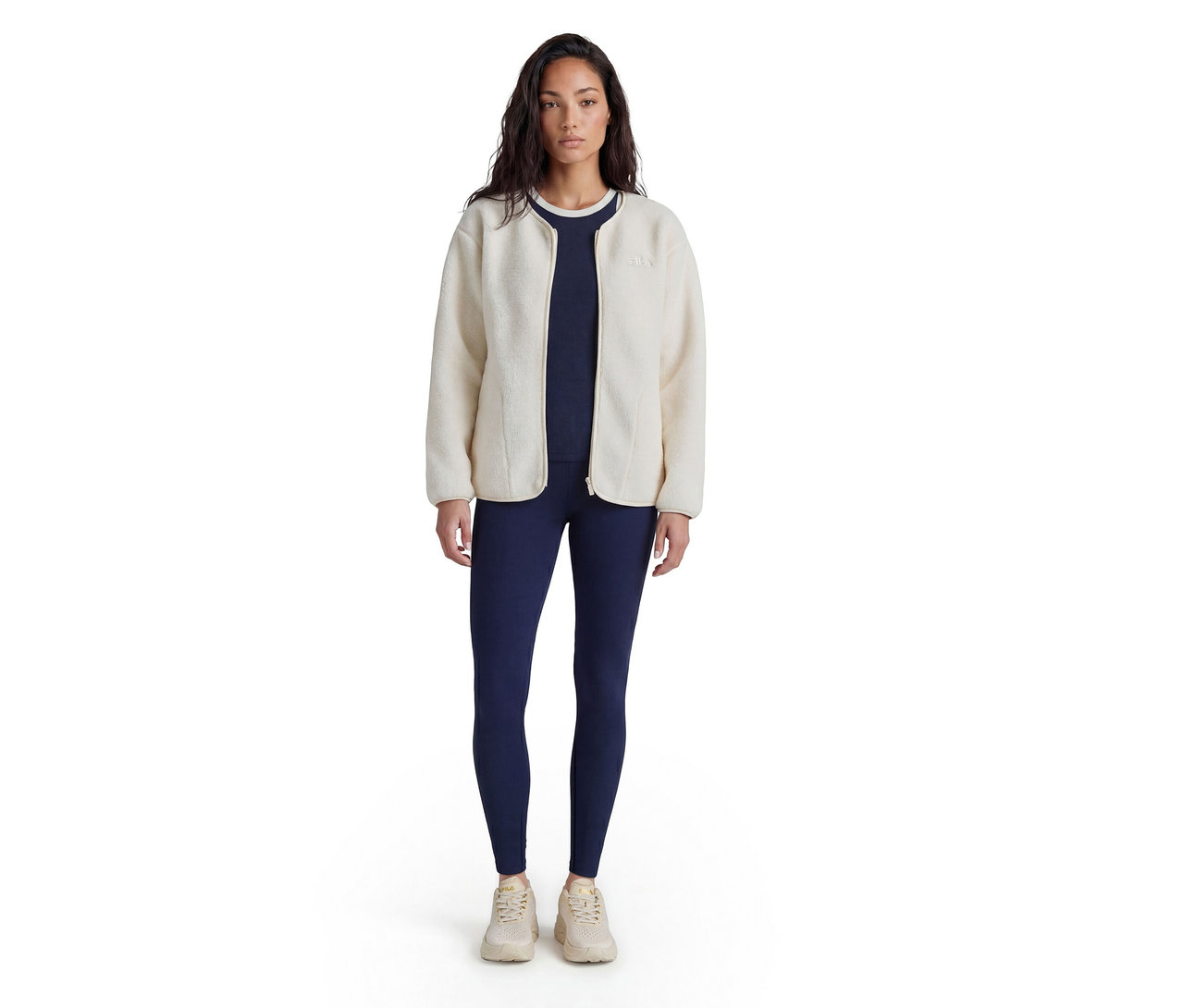 Femme debout, portant une veste polaire crème sur un haut et un legging bleu marine, avec des baskets claires.