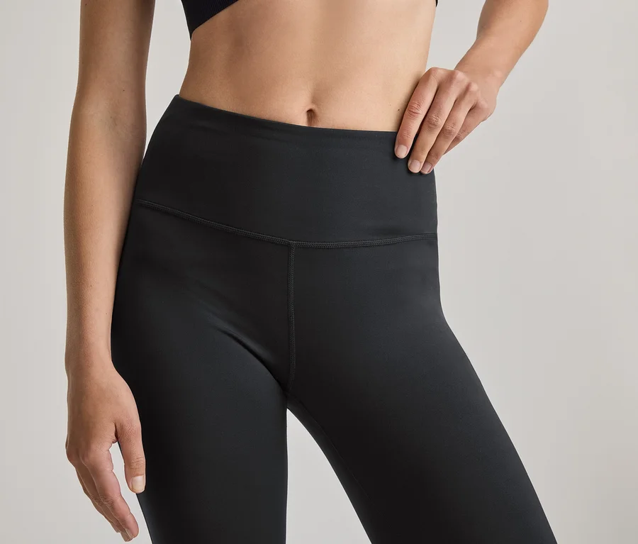 Gros plan sur le torse d'une personne portant un legging noir taille haute et une brassière de sport noire. Sa main droite est posée sur sa hanche.