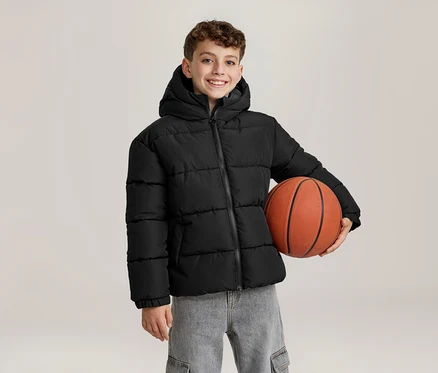 Un jeune garçon souriant vêtu d'une doudoune noire à capuche et d'un jean gris tient un ballon de basket orange sous son bras gauche.