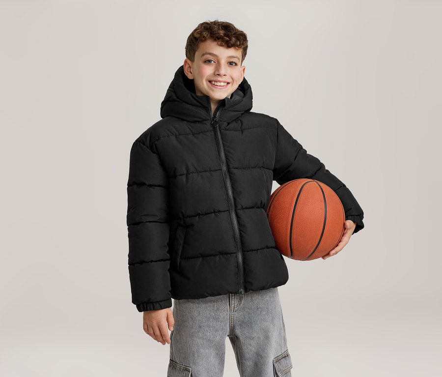Un jeune garçon souriant vêtu d'une doudoune noire à capuche et d'un jean gris tient un ballon de basket orange sous son bras gauche.