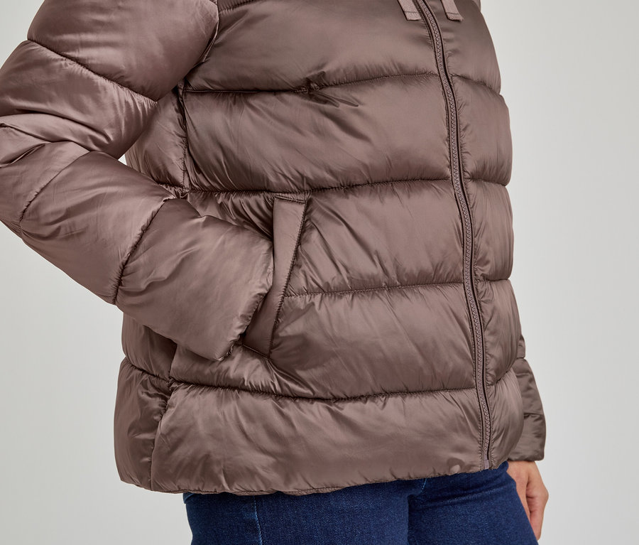 Gros plan d'une personne portant une doudoune marron et un jean bleu, la main gauche dans la poche de la veste.