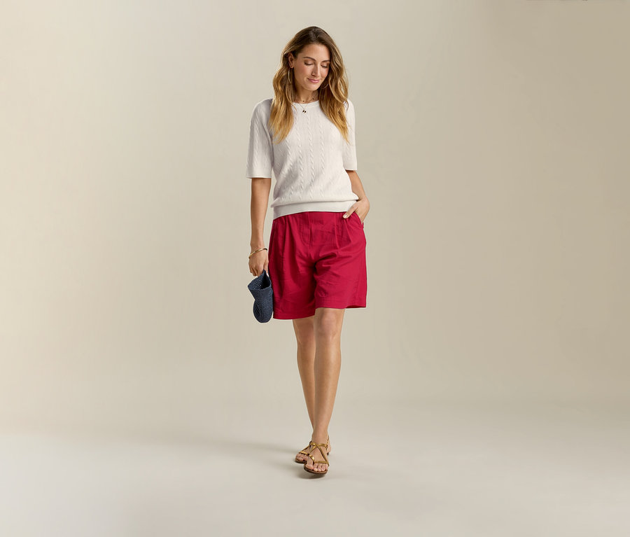 Femme en pull blanc et bermuda en lin rouge, une main dans la poche, tenant une visière bleue.