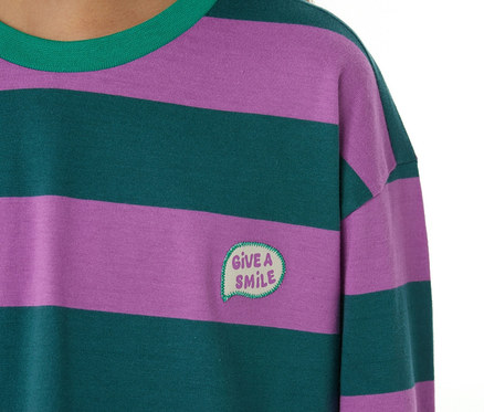 Gros plan d'un enfant portant un pyjama. Le haut de pyjama a des rayures horizontales violettes et vert foncé, un col vert et une bulle de dialogue brodée avec "GIVE A SMILE" sur la poitrine.