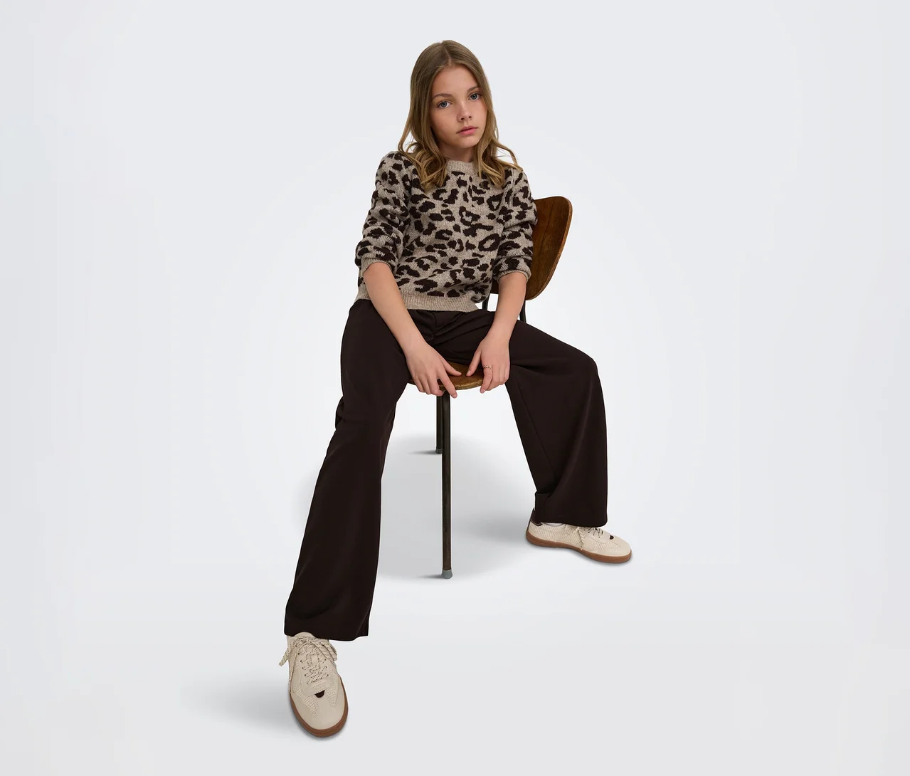 Jeune fille assise sur une chaise en bois, portant un pull à imprimé léopard, un pantalon large marron foncé et des baskets claires.