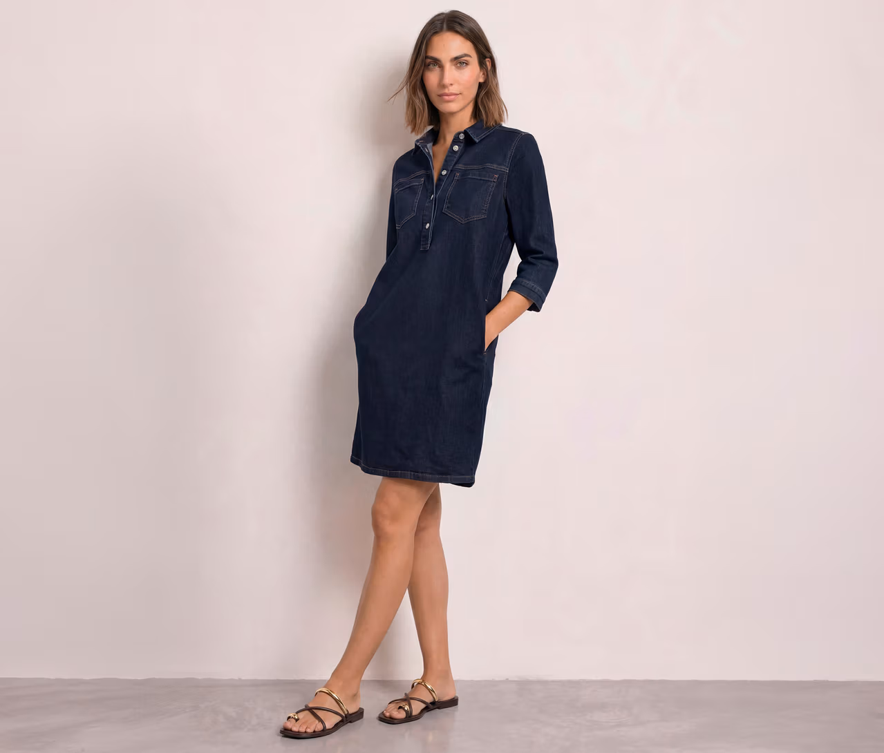 Femme portant une robe en jean Street One bleu foncé avec col, poches poitrine et manches 3/4, les mains dans les poches. Elle porte des sandales foncées.