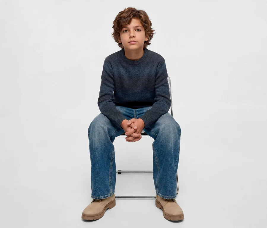 Un garçon aux cheveux bouclés est assis sur une chaise, portant un pull dégradé bleu foncé, un jean bleu et des bottes marron clair, regardant directement l'appareil photo.
