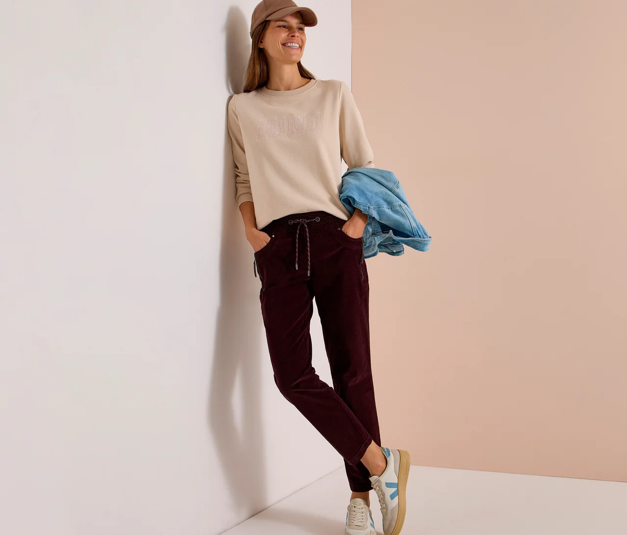 Femme souriante portant une casquette beige, un haut à manches longues beige et un pantalon en velours côtelé CECIL rouge foncé, appuyée contre un mur avec une veste en jean sur le bras.