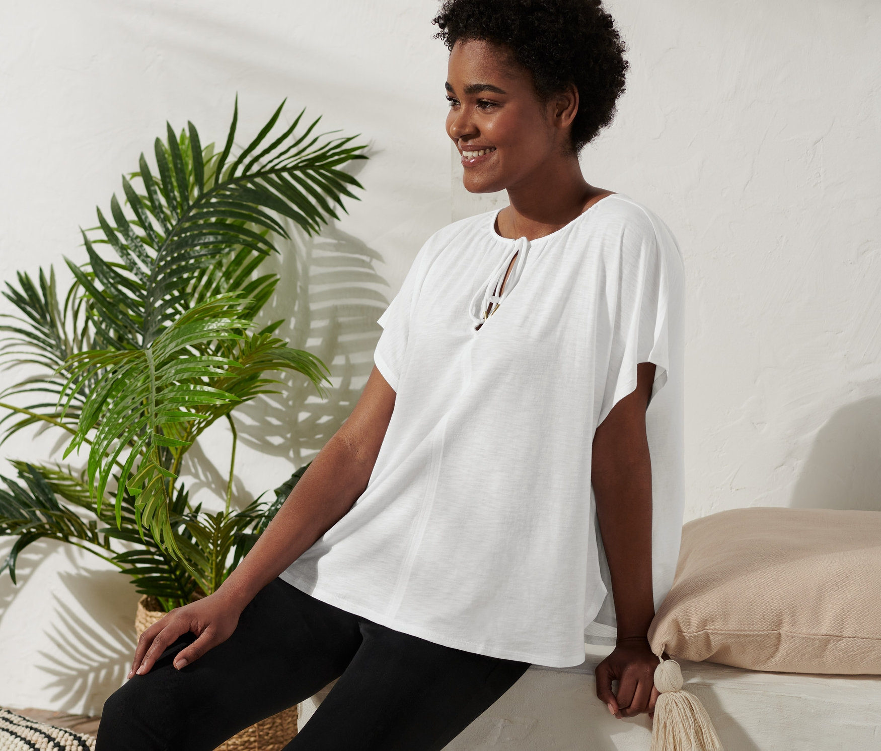 Une femme est assise et sourit, elle porte une blouse avec lien à nouer, un pantalon noir et des sandales compensées.