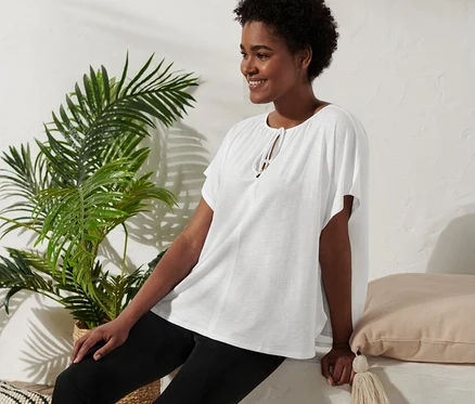 Une femme est assise et sourit, elle porte une blouse avec lien à nouer, un pantalon noir et des sandales compensées.