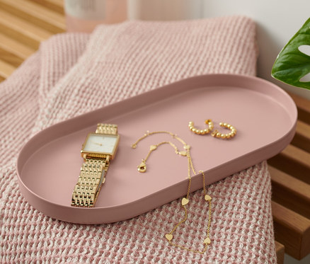 Sur un banc en bois se trouvent un plateau en métal couleur rouille, une montre, un collier et des boucles d'oreilles. À côté du plateau se trouvent une serviette rose et une bouteille avec un distributeur.