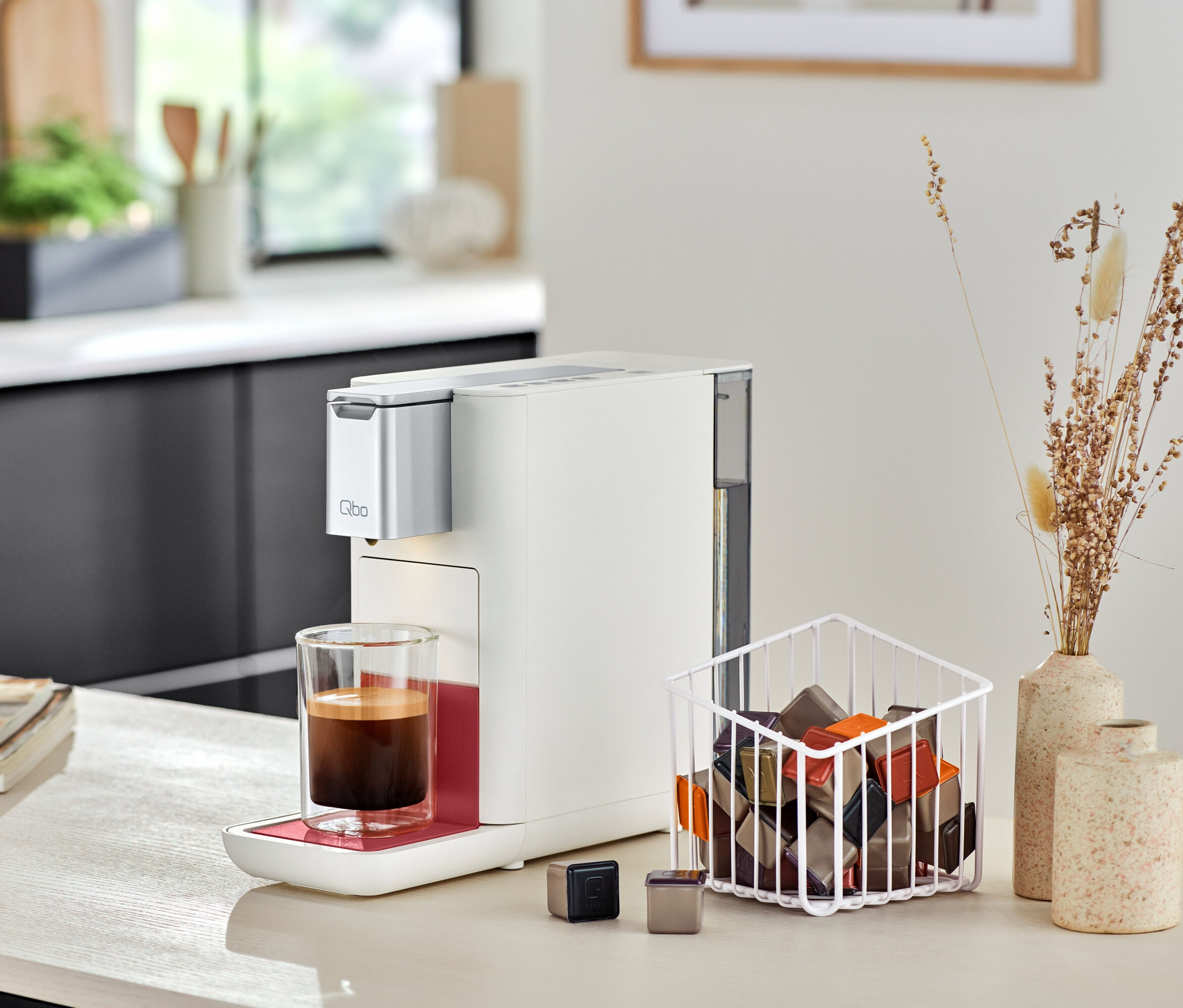 Machine à café Qbo ICONIC Dark Chrome à côté de Qbo ESSENTIAL Bright Coral avec tasse de café et capsules.