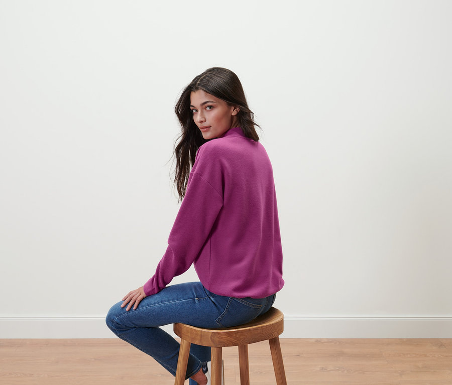 Une femme est assise sur un tabouret et porte un sweat-shirt violet avec un col montant et un jean.