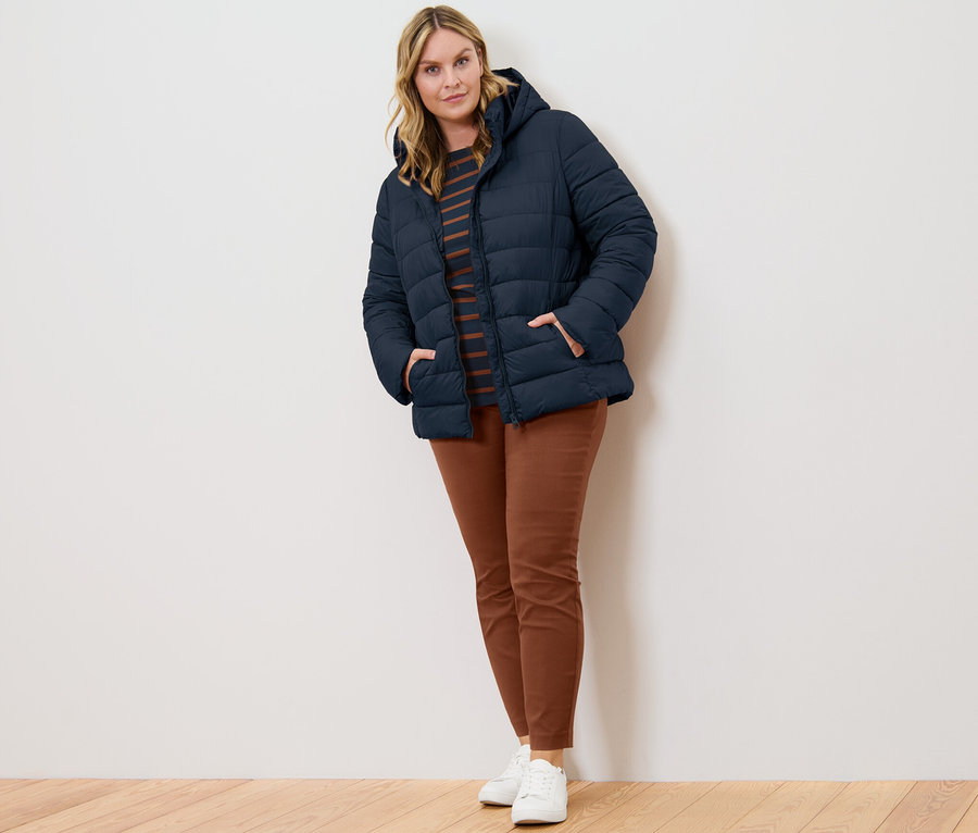 Femme posant en veste matelassée à capuche bleu foncé, t-shirt manches 3/4 à rayures et pantalon stretch cognac.