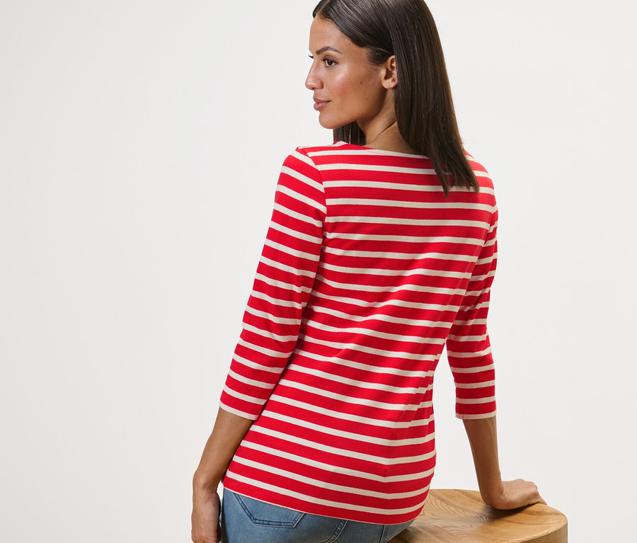Femme assise de côté sur un tabouret en bois portant un t-shirt rayé manches 3/4, à rayures rouge.