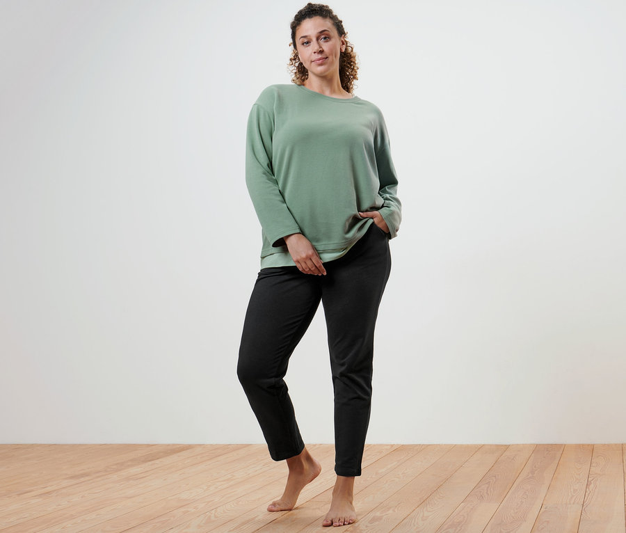 Une femme pose dans un haut à manches longues fitness oversize et un pantalon de jogging noir.