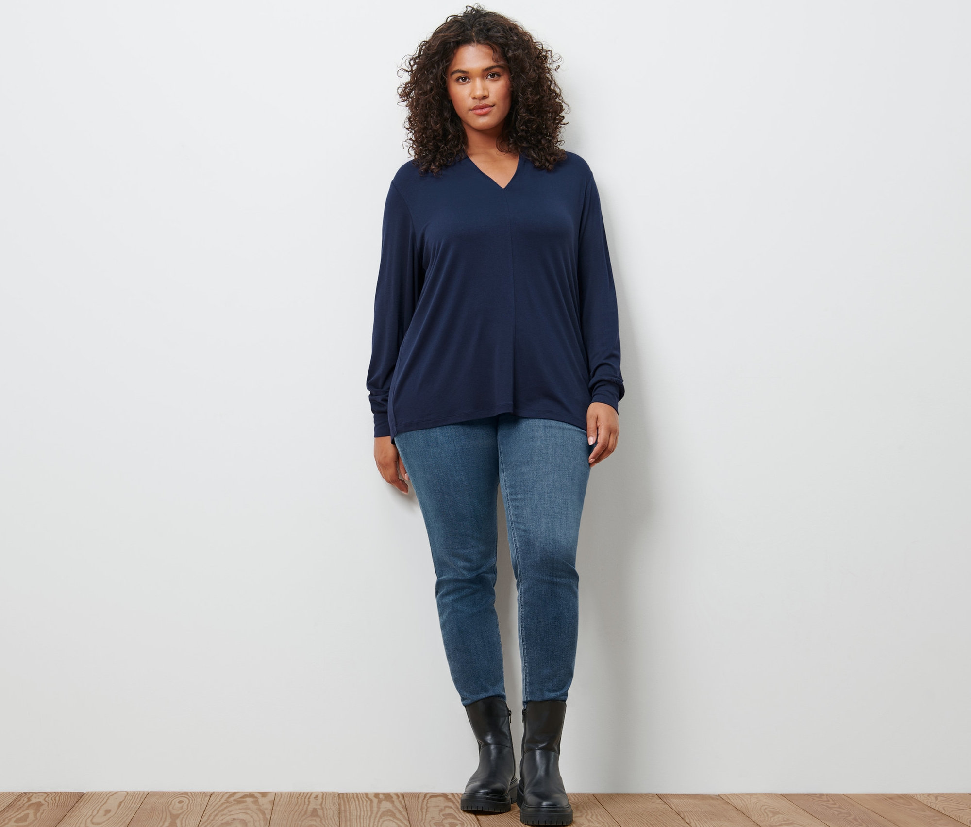 Une femme pose portant un t-shirt manches longues avec encolure en V bleu foncé, un jean slim en denim « Fit Emma » bleu moyen et des bottines en cuir.