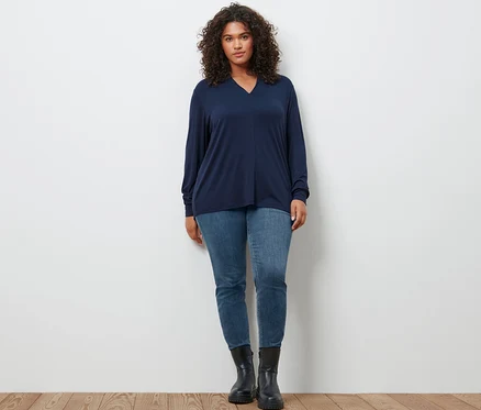 Une femme pose portant un t-shirt manches longues avec encolure en V bleu foncé, un jean slim en denim « Fit Emma » bleu moyen et des bottines en cuir.
