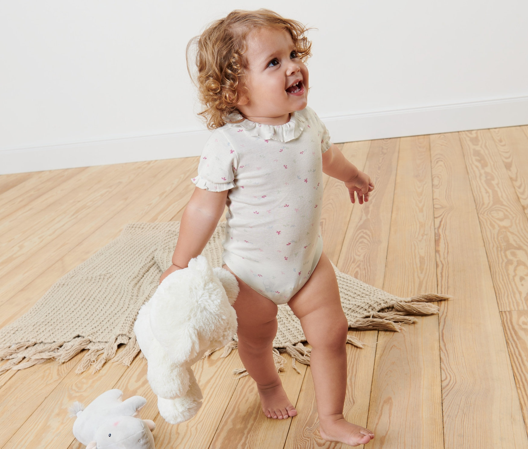Un jeune enfant se tient sur un plancher en bois et tient des peluches. Il porte 2 bodys pour bébé.