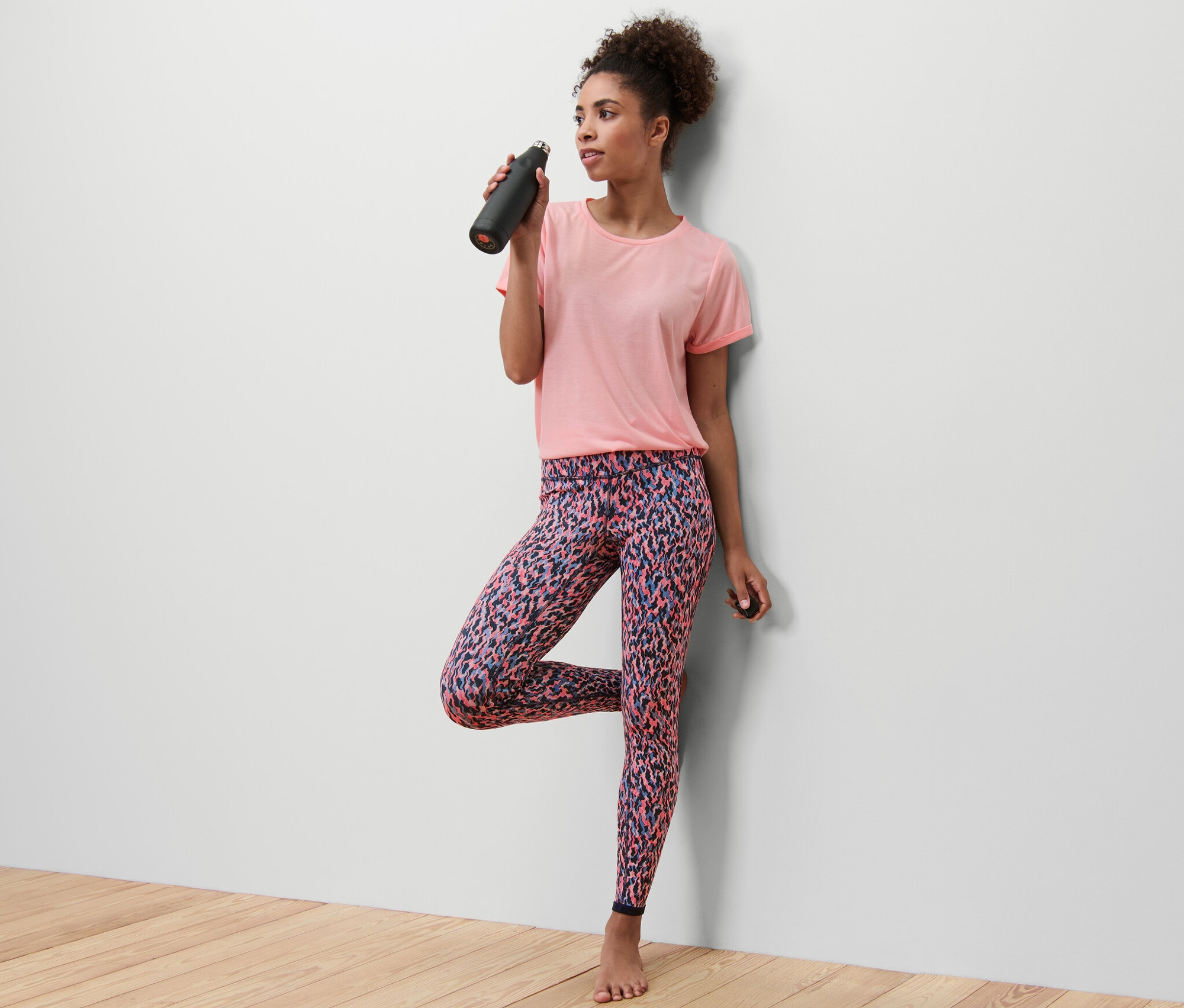 Femme en t-shirt rose en équilibre sur une jambe, buvant dans une bouteille noire. Elle porte un legging de sport réversible, rose/bleu marine.