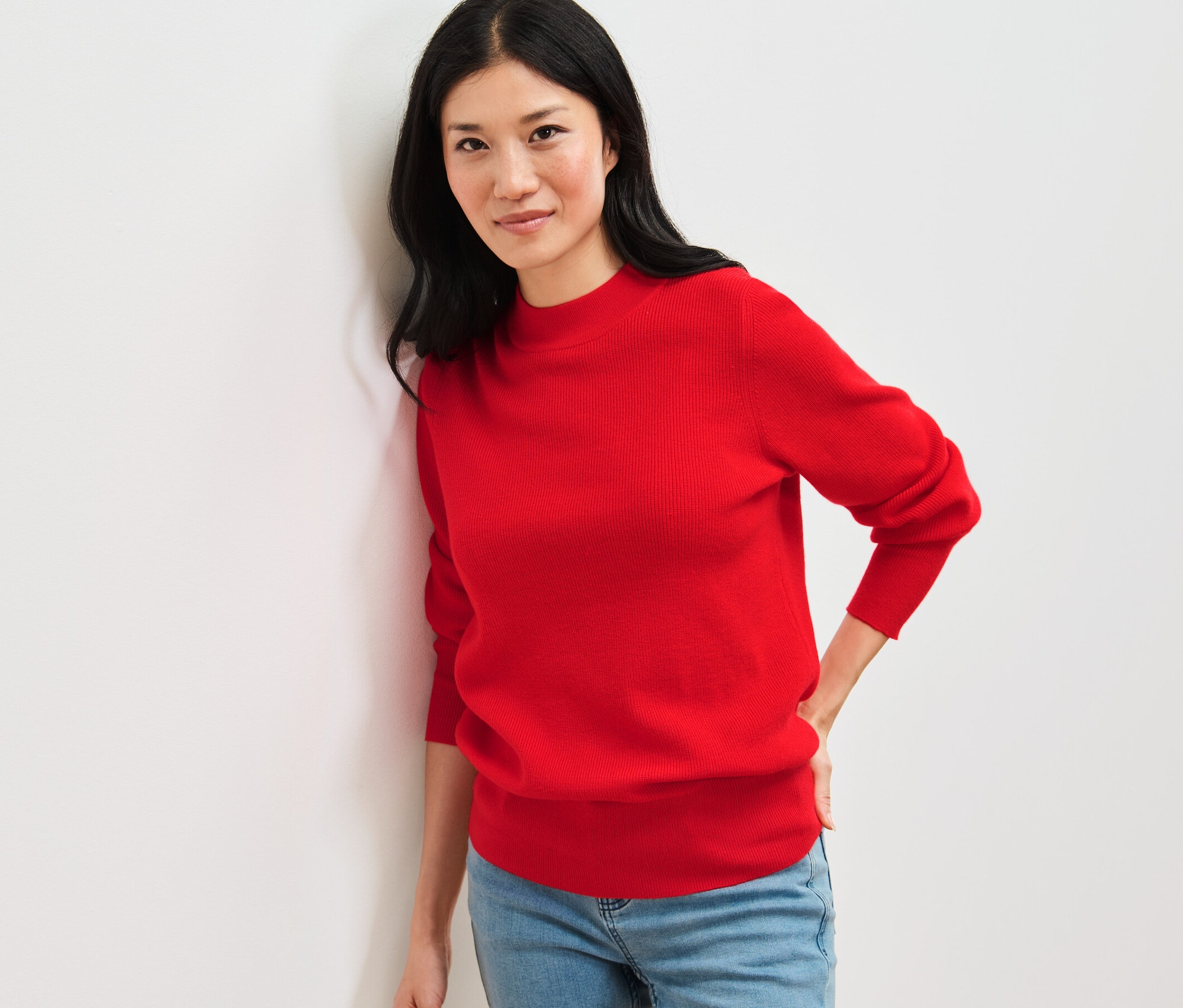 Une femme appuyée contre un mur portant un pull en maille fine rouge et un jean.