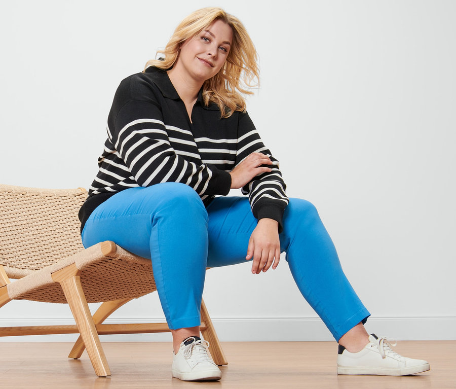 Une femme blonde est assise sur un fauteuil en osier et porte un pull avec col, un pantalon en bengaline et des baskets en cuir.