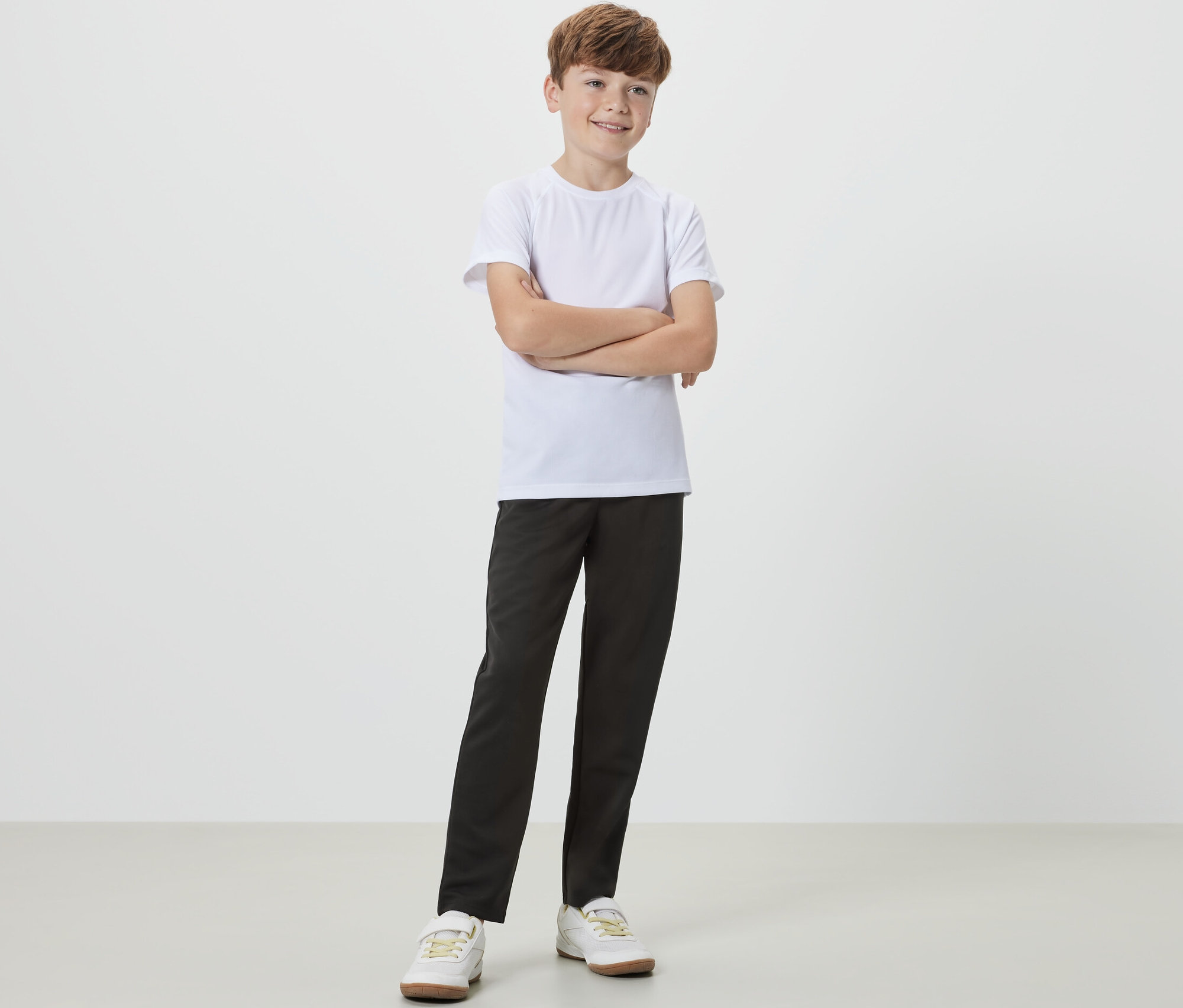 Un garçon se tient les bras croisés et porte un pantalon de sport noir pour enfant et un t-shirt blanc.