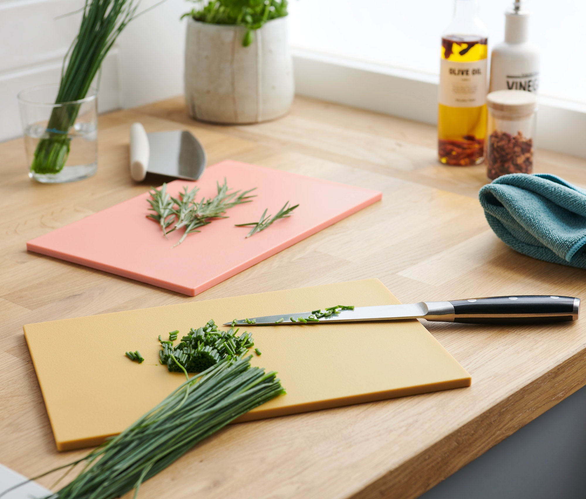 Deux planches à découper sur un comptoir en bois : une jaune avec de la ciboulette hachée et un couteau, une rose avec du romarin.