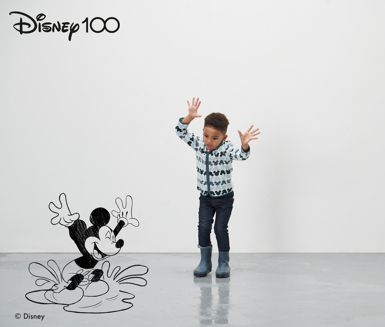 Un garçon se tient devant un personnage de Mickey Mouse dessiné et agite les mains. Il porte une veste en polaire bleue pour tout-petit « Mickey ».