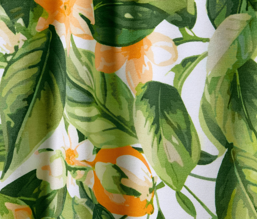 Gros plan sur un pantalon palazzo imprimé avec des feuilles et des fleurs dans les tons de vert et d'orange sur un fond blanc.