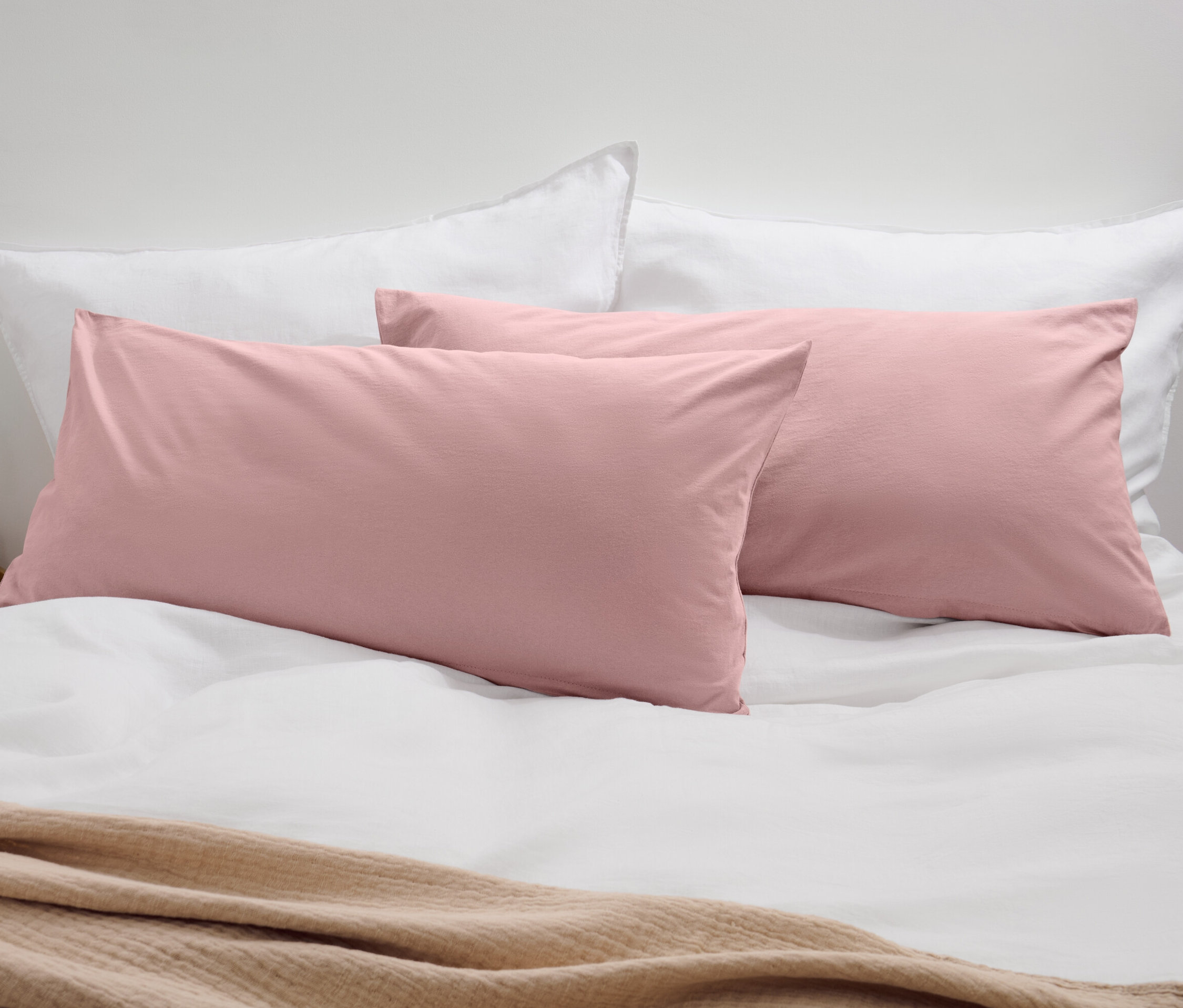 Deux taies d’oreiller en jersey rose sur un lit avec des oreillers blancs et une couverture beige.