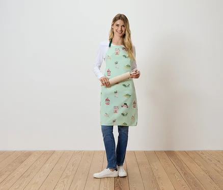 Une femme debout tient un rouleau à pâtisserie. Elle porte un tablier vert clair avec un imprimé.