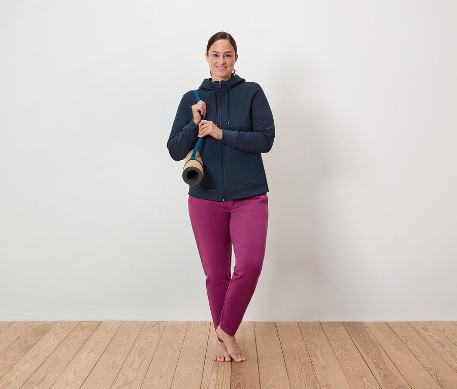 Une femme se tient sur un plancher en bois, un tapis de yoga sur l'épaule, et porte un sweat-shirt bleu à fermeture éclair et un pantalon violet.