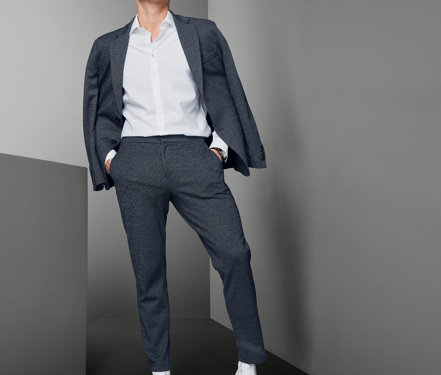 Un homme pose en veste grise, chemise blanche, pantalon en jersey et baskets blanches.