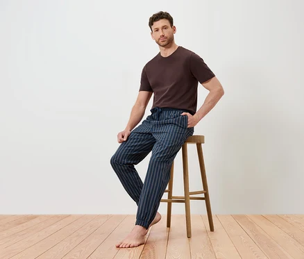Un homme est assis sur un tabouret en bois, portant un t-shirt marron et un pantalon rayé bleu.