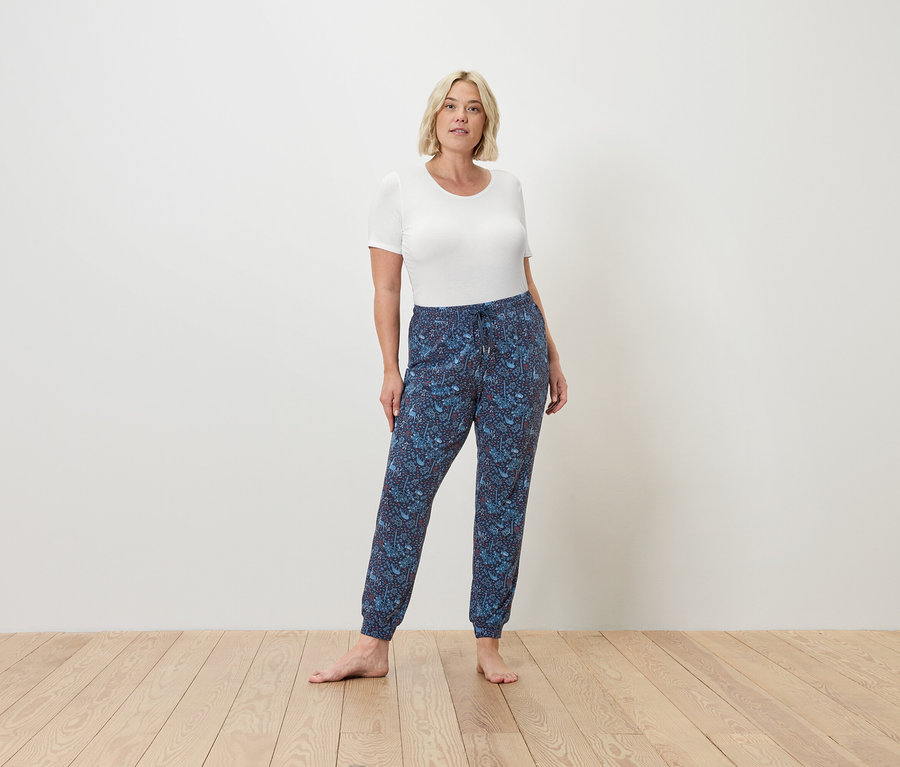 Une femme se tient pieds nus sur un plancher en bois, portant un t-shirt blanc et un pantalon de jogging bleu.