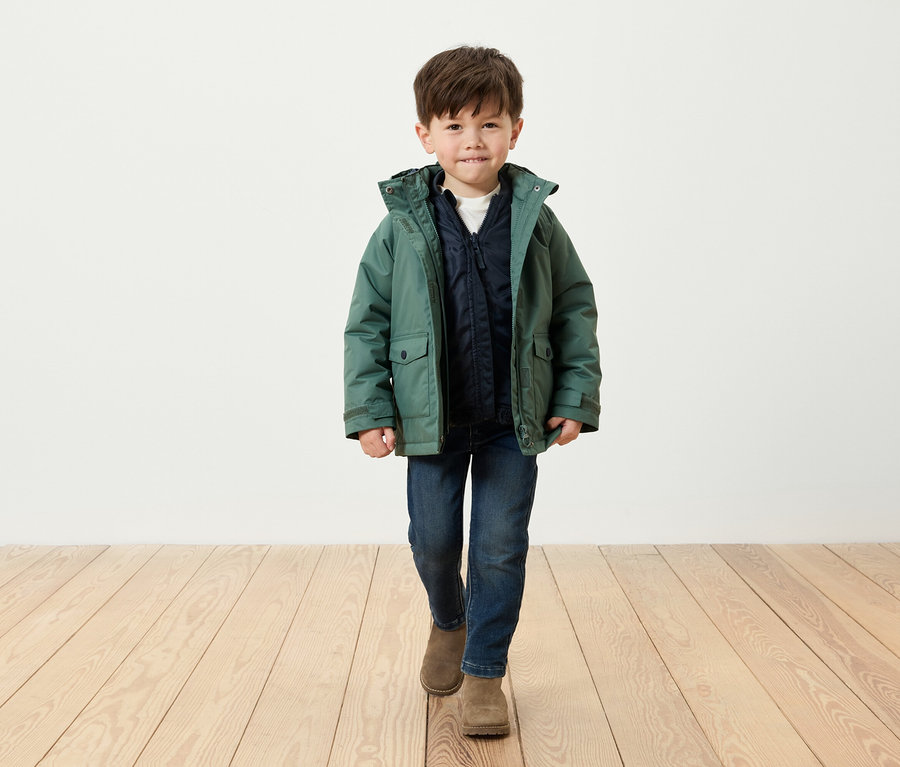Un garçon vêtu d'une veste verte, d'un jean bleu et de bottes marron marche sur un plancher en bois.