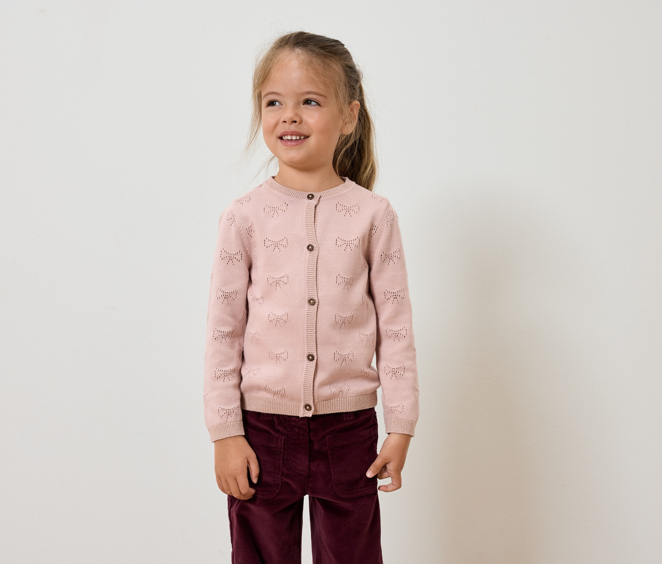 Une fille aux cheveux bruns porte un cardigan rose et un pantalon violet.