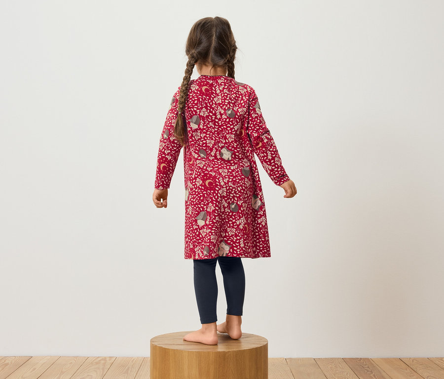 Une fille portant une robe rouge à motifs et des leggings bleu marine se tient sur un socle en bois.