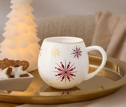 Sur un plateau doré se trouvent une tasse blanche avec des flocons de neige rouges et dorés, un bol de pain d'épices et un arbre lumineux.
