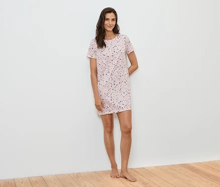Une femme se tient devant un mur blanc, portant une chemise de nuit rose à manches courtes avec des points noirs.