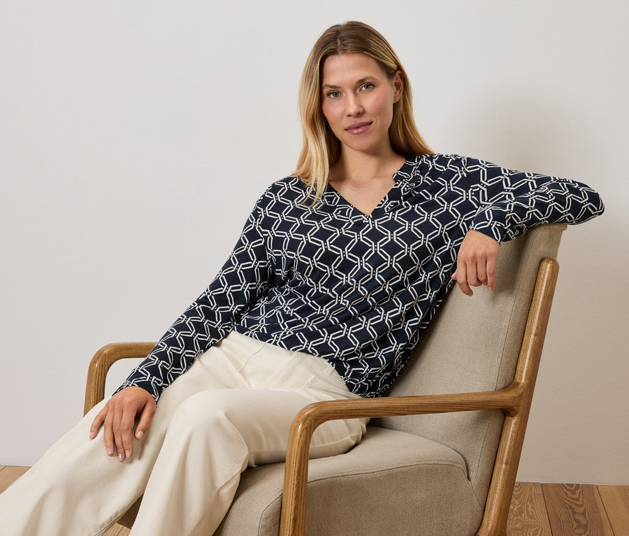 Une femme est assise dans un fauteuil et porte un haut bleu et blanc et un pantalon crème.