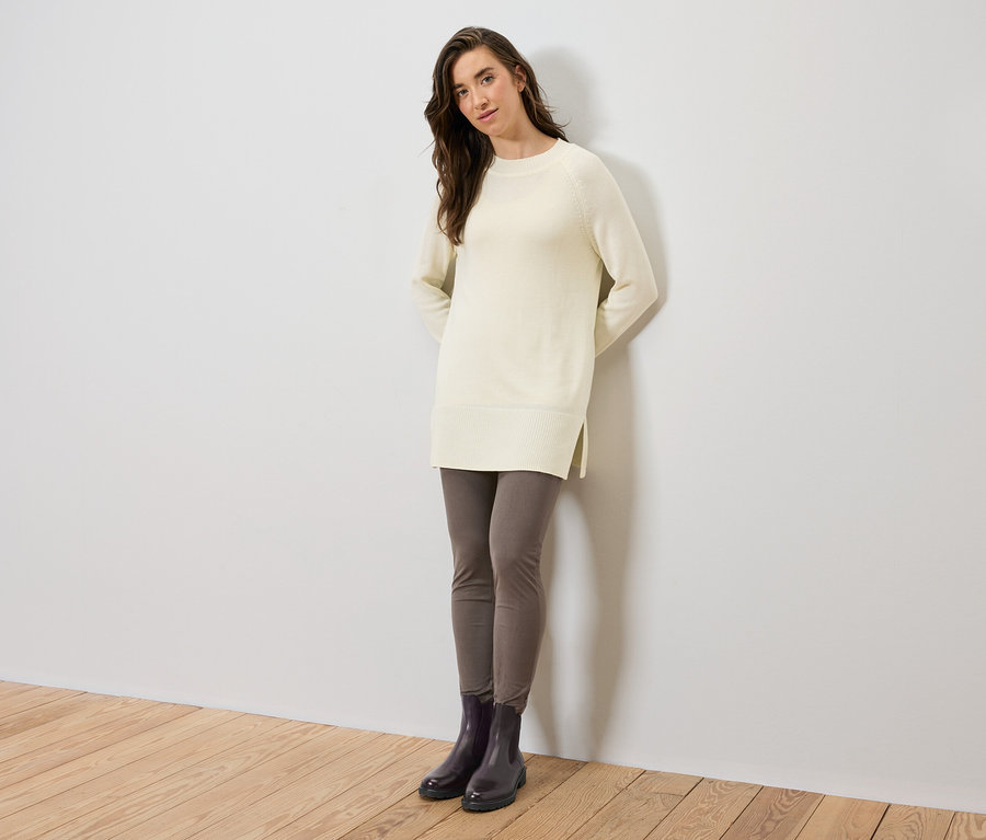 Une femme debout porte un long pull en tricot crème, des leggings gris et des bottes noires.