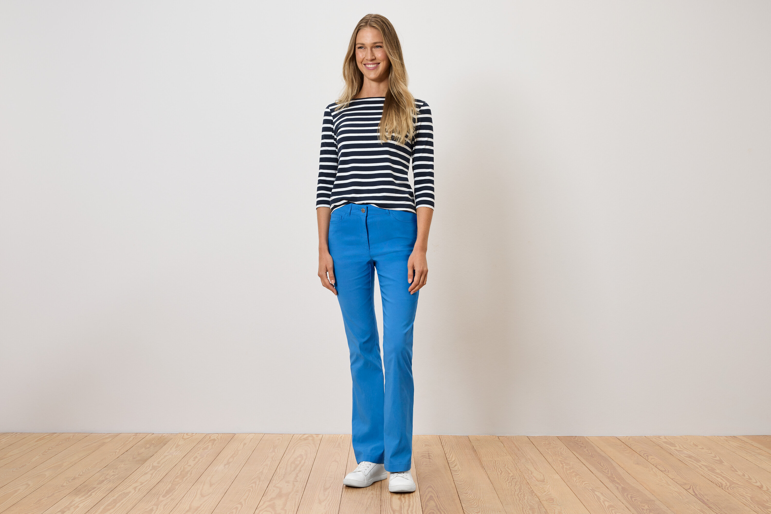 Une femme debout porte un pantalon bleu, un t-shirt rayé et des chaussures blanches.