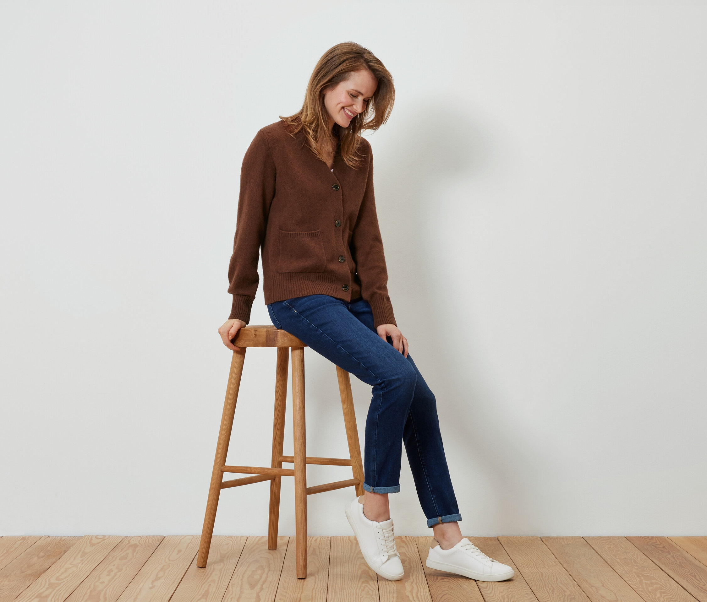 Une femme est assise sur un tabouret en bois, portant un cardigan marron, un jean et des baskets blanches.