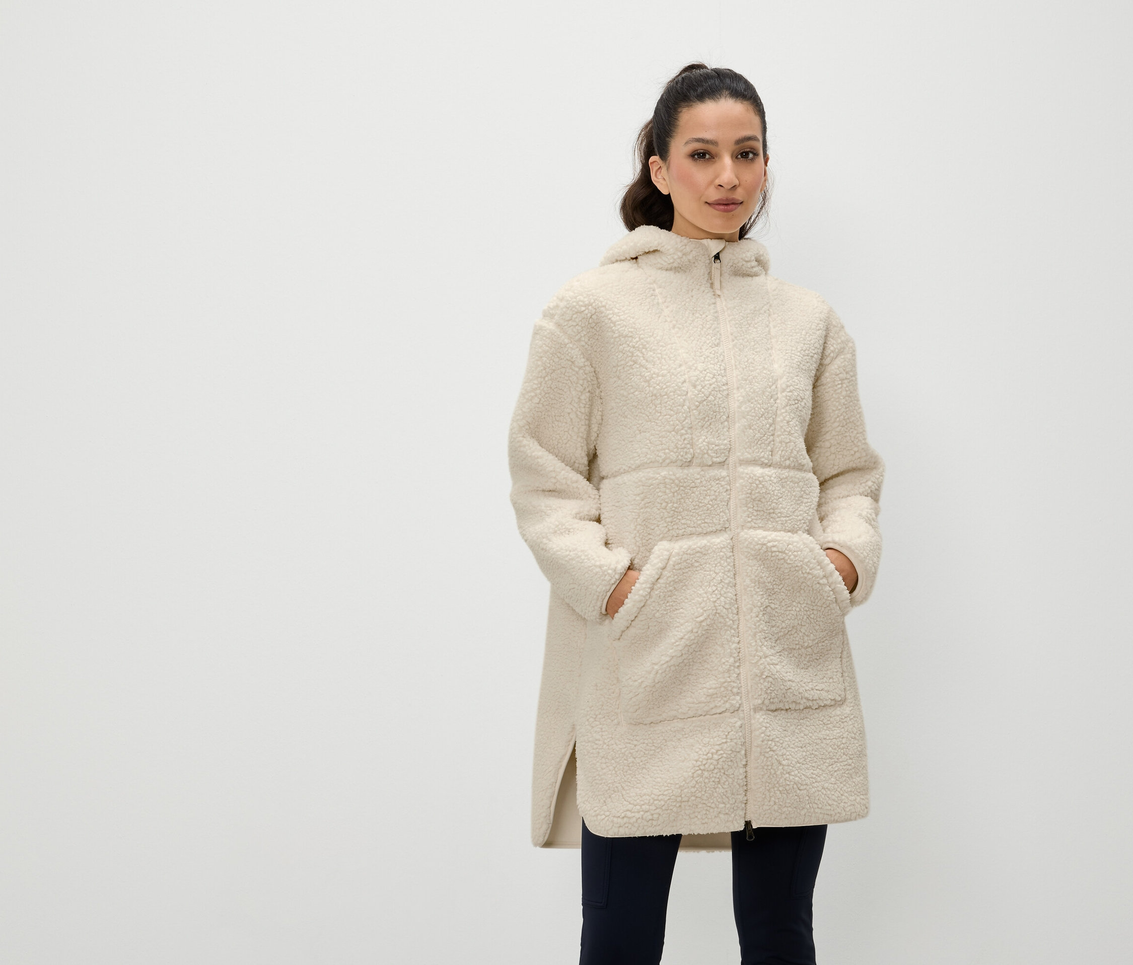 Une femme se tient debout, les mains dans les poches d'un manteau crème zippé à capuche.
