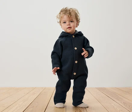 Un enfant aux cheveux bouclés se tient sur un plancher en bois, portant une combinaison bleu foncé à capuche et boutonnée.